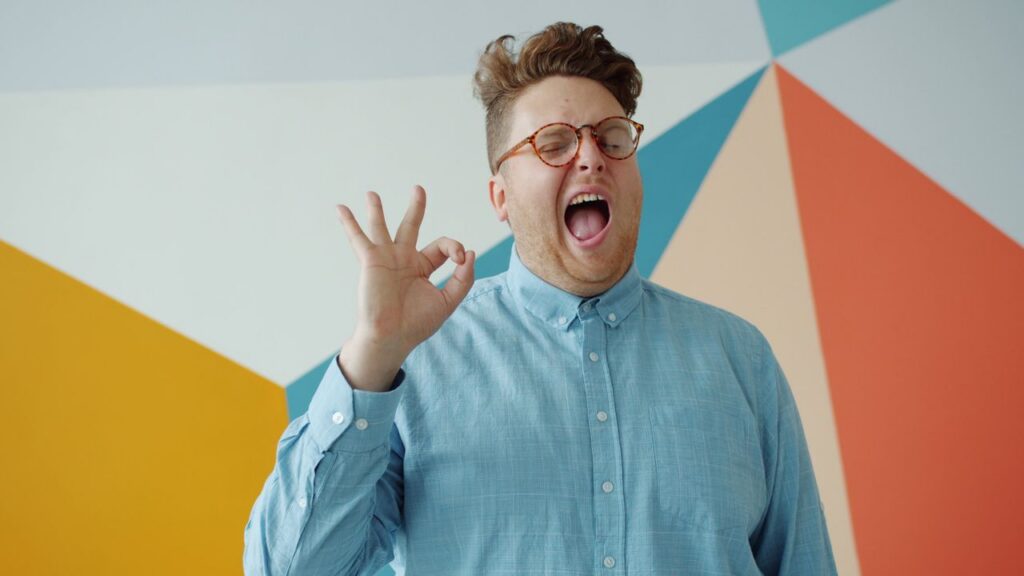 Smiling man in blue shirt making an okay sign against colorful wall.